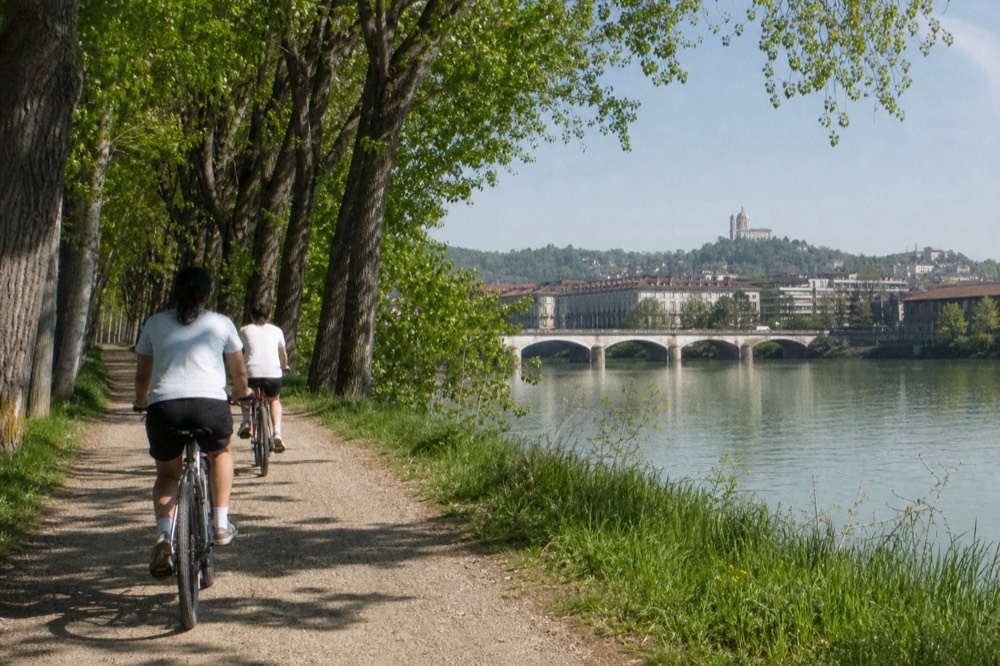 Cyclists on a tree-lined path along the Po river in Turin with the hill of Superga behind.