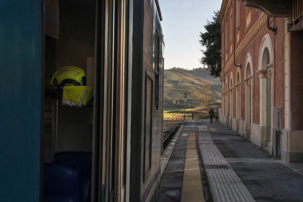 Regional train at a small Italian station platform with hills in the background.