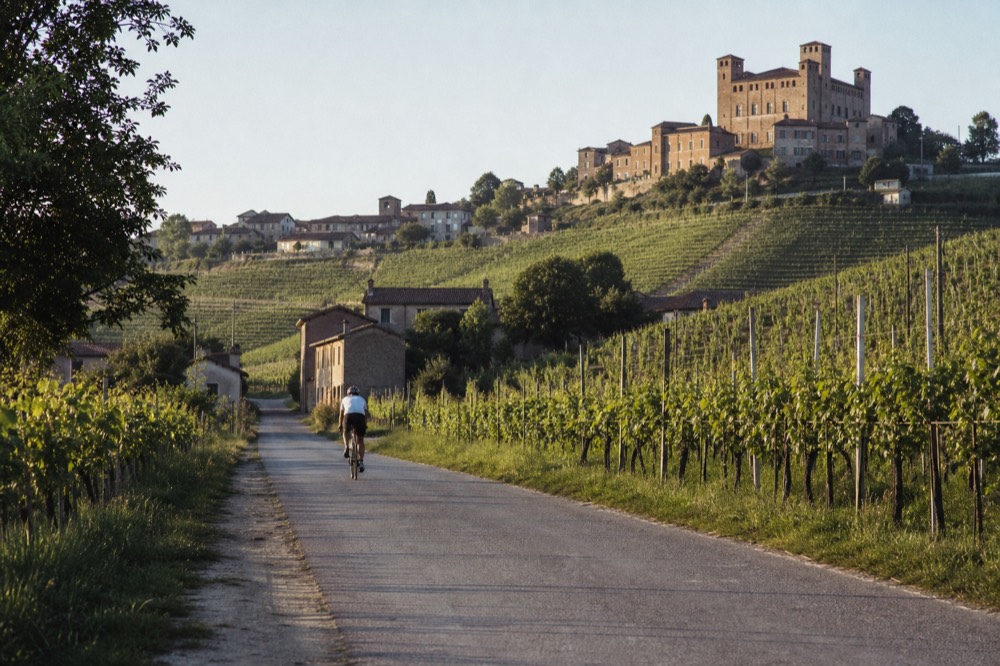 A cyclist on a quiet Langhe road between vineyard rows with hilltop village silhouettes.